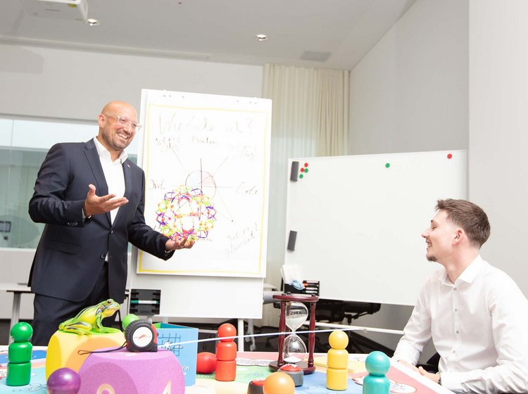 Ein Mann im Anzug erklärt an einem Flipchart ein buntes Diagramm, während zwei Personen ihm aufmerksam zuhören. Auf dem Tisch vor ihnen liegen große bunte Spielfiguren und ein Würfel, die für eine spielerische Präsentation oder Schulung genutzt werden.