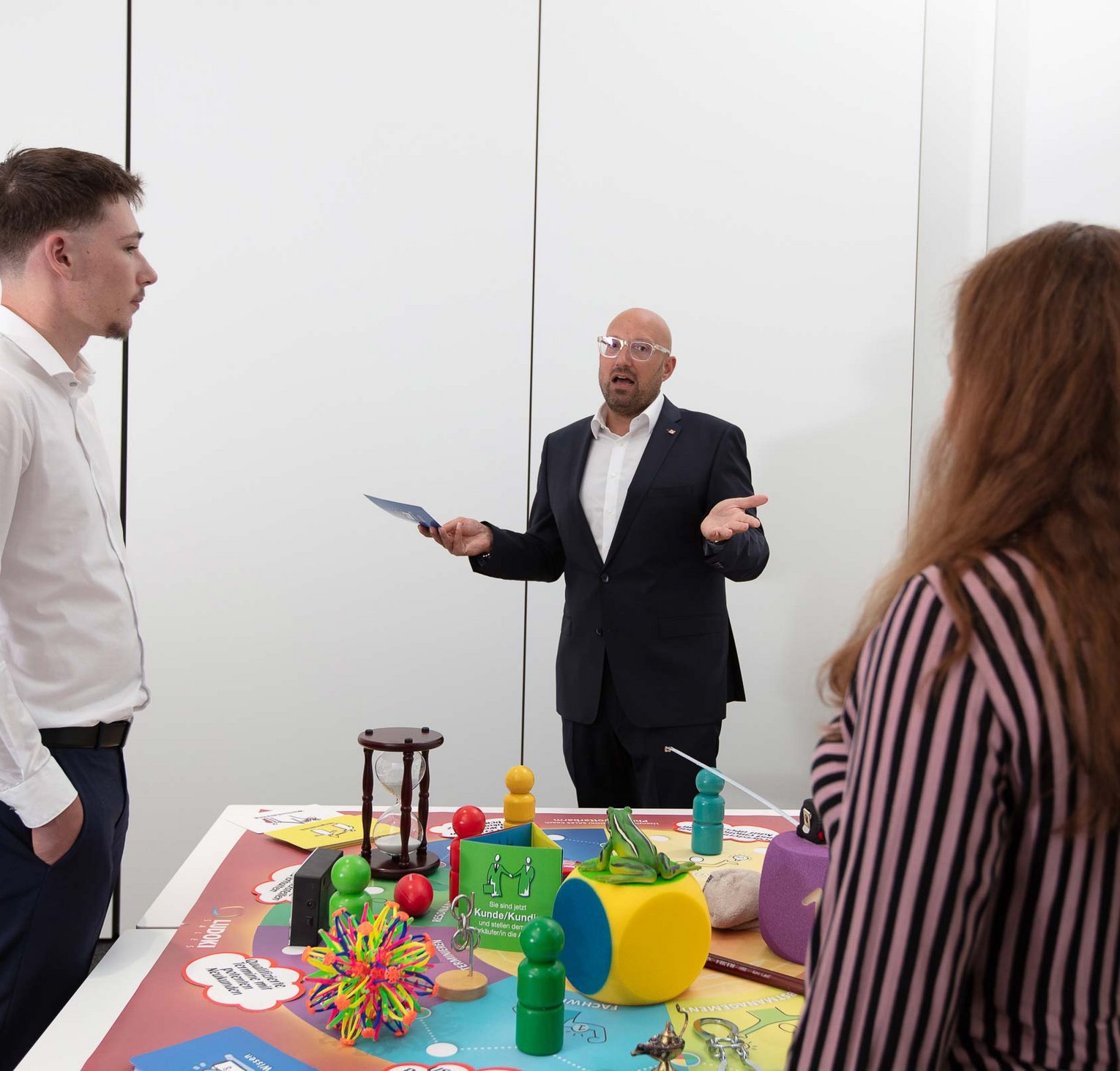 Drei Personen stehen um einen Tisch mit bunten Spiel- und Lernmaterialien. Ein Mann im Anzug spricht deutlich und erklärt etwas, während die anderen beiden aufmerksam zuhören. Die Szene wirkt wie eine Präsentation oder Schulung.