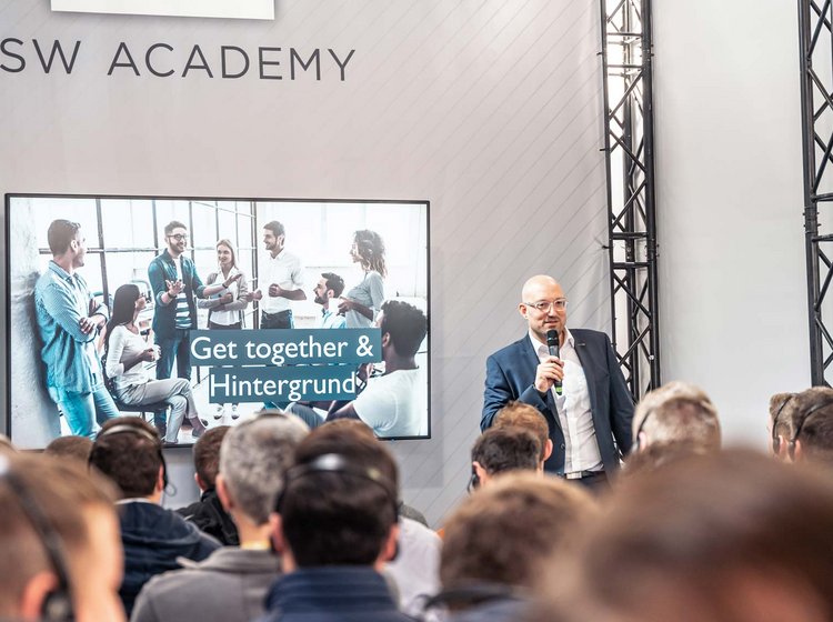 Ein Mann hält eine Präsentation vor einem Publikum in einem Seminarraum der PSW Academy. Auf einem Bildschirm im Hintergrund ist eine Gruppe von Menschen zu sehen, darunter der Text „Get together & Hintergrund“.