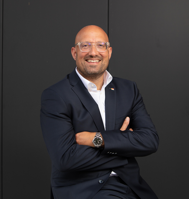 Porträt eines lächelnden Mannes mit Brille, Anzug und verschränkten Armen vor dunklem Hintergrund.