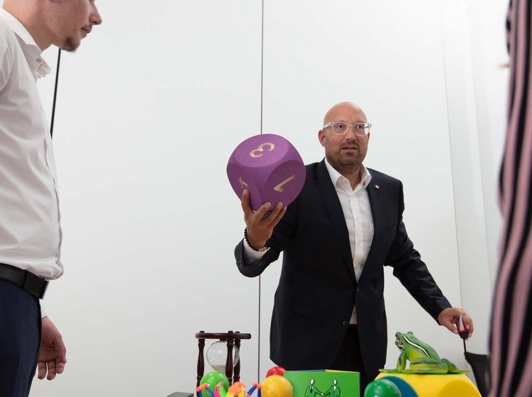 Zwei Männer stehen an einem Tisch mit verschiedenen bunten Objekten, einer von ihnen hält einen großen, lila Würfel mit Zahlen in der Hand, offenbar in einer Diskussion oder Präsentation.