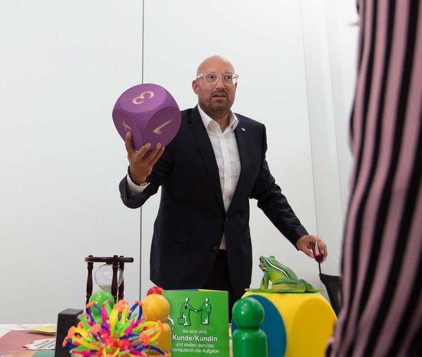 Zwei Männer stehen an einem Tisch mit verschiedenen bunten Objekten, einer von ihnen hält einen großen, lila Würfel mit Zahlen in der Hand, offenbar in einer Diskussion oder Präsentation.