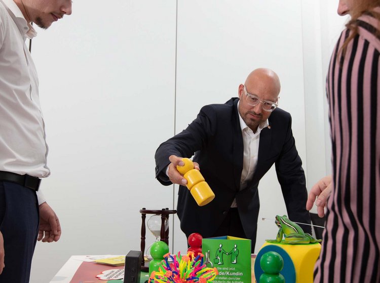 Drei Menschen stehen um einen Tisch mit verschiedenen bunten Spiel- und Lernmaterialien, ein Mann gießt Wasser aus einer gelben Gießkanne auf die Gegenstände.