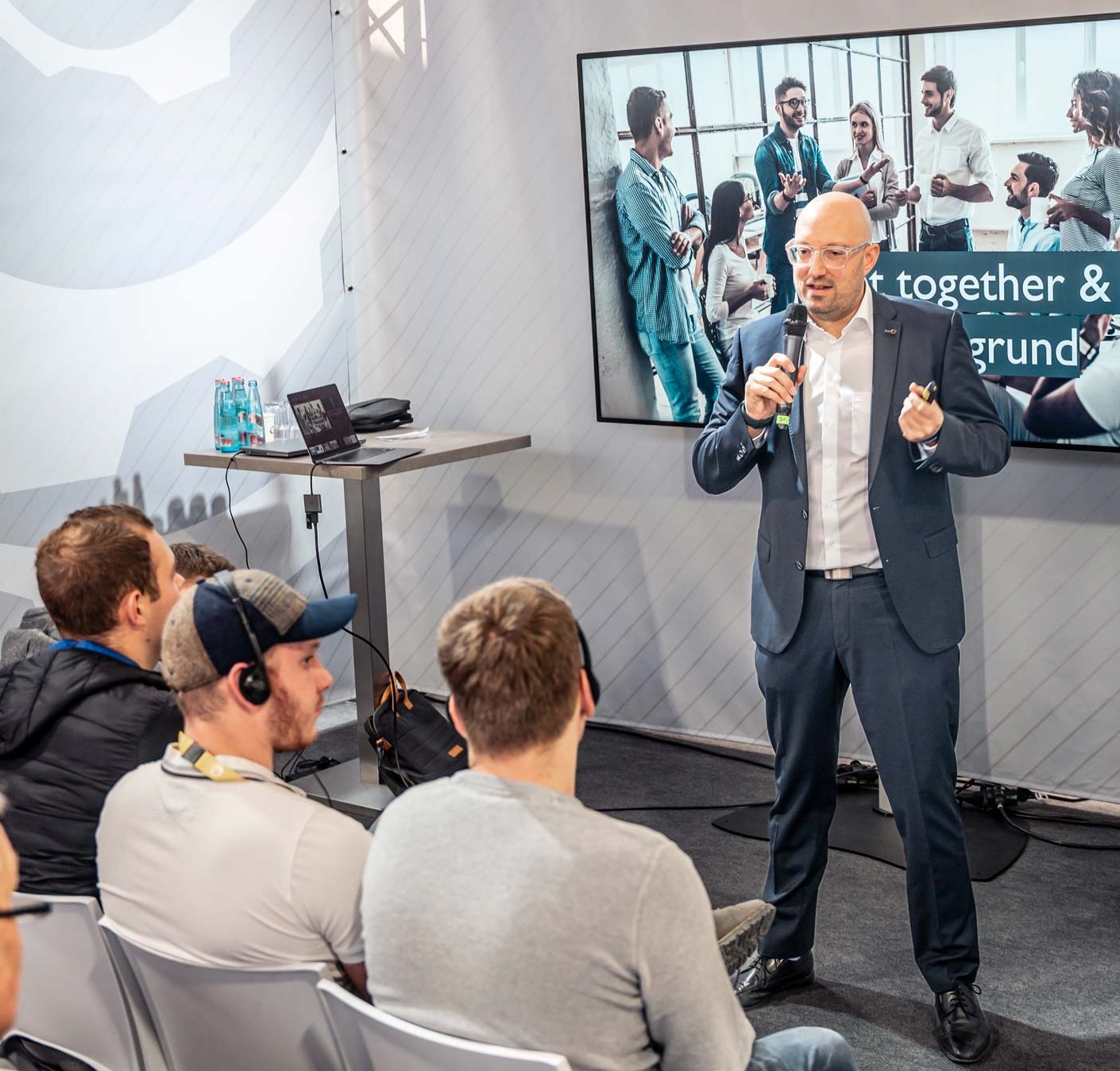 Ein Mann im Anzug hält eine Präsentation vor einer Gruppe Zuhörer, die ihn aufmerksam ansehen. Im Hintergrund zeigt ein Bildschirm eine Teamgruppe mit dem Text „Together & ...“. Die Atmosphäre wirkt professionell und konzentriert.