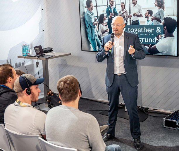 Ein Mann im Anzug hält eine Präsentation vor einer Gruppe Zuhörer, die ihn aufmerksam ansehen. Im Hintergrund zeigt ein Bildschirm eine Teamgruppe mit dem Text „Together & ...“. Die Atmosphäre wirkt professionell und konzentriert.