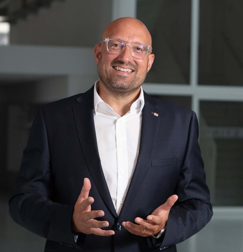 Ein lächelnder Mann in Anzug und Brille steht in einem hellen, modernen Innenraum und gestikuliert freundlich mit den Händen.