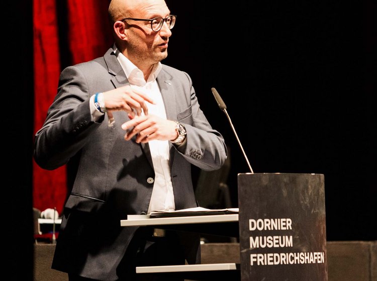 Ein Mann in Anzug hält eine Rede oder Präsentation am Rednerpult mit der Aufschrift „Dornier Museum Friedrichshafen“ vor dunklem Hintergrund mit roter Beleuchtung.