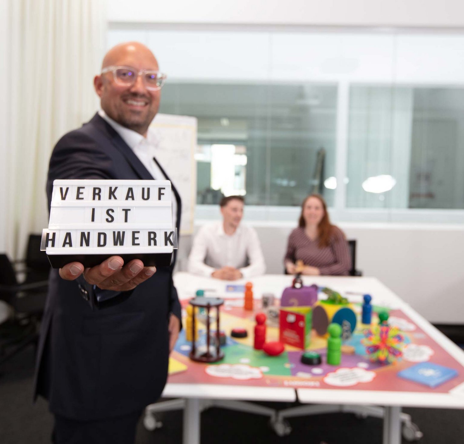Ein Mann hält ein Schild mit der Aufschrift „Verkauf ist Handwerk“. Im Hintergrund sitzen zwei Personen an einem Tisch mit bunten Spielzeugen und Lernmaterialien.
