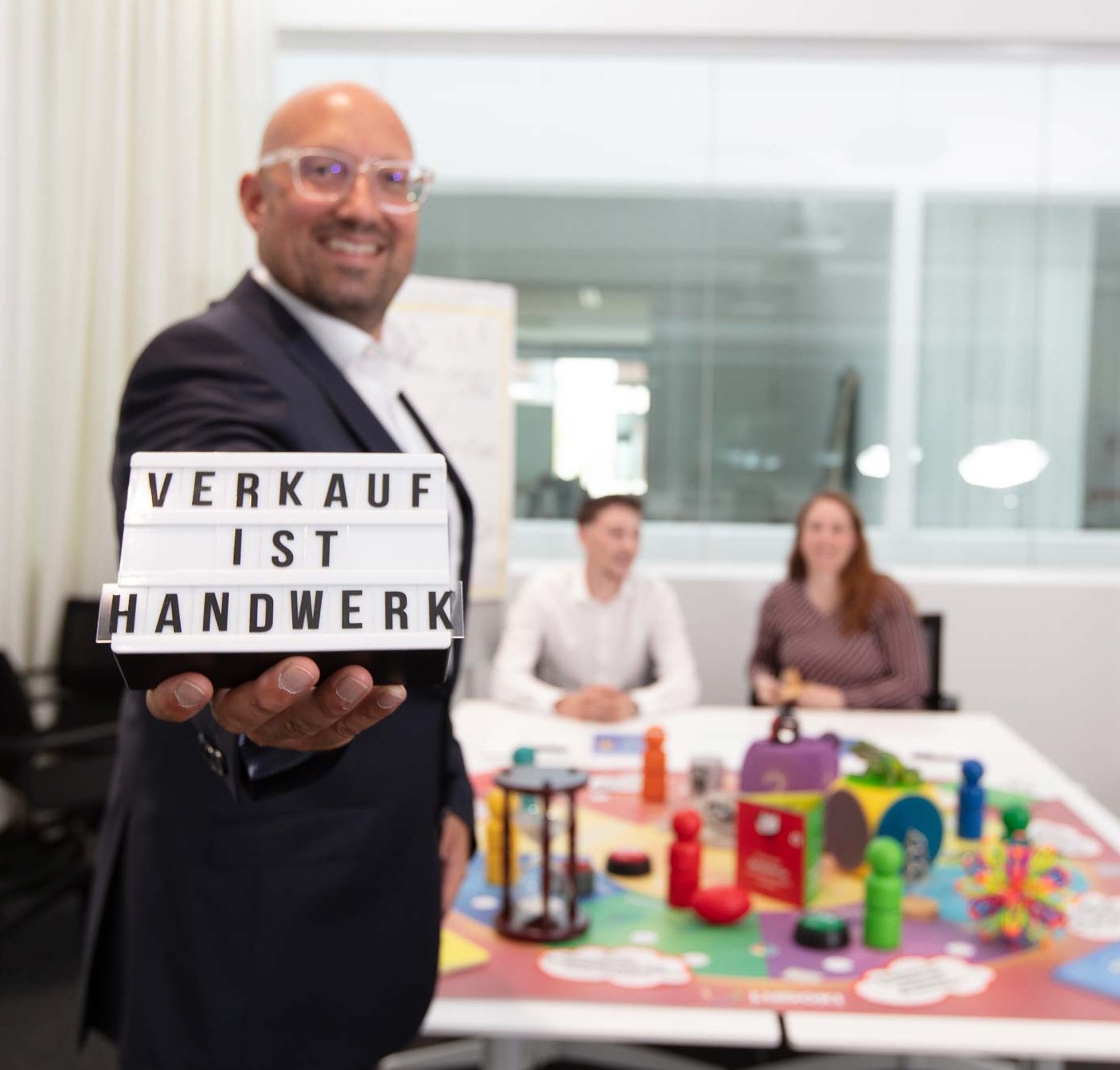 Ein Mann hält ein Schild mit der Aufschrift „Verkauf ist Handwerk“. Im Hintergrund sitzen zwei Personen an einem Tisch mit bunten Spielzeugen und Lernmaterialien.