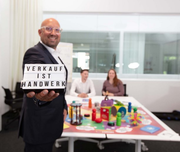 Ein Mann hält ein Schild mit der Aufschrift „Verkauf ist Handwerk“. Im Hintergrund sitzen zwei Personen an einem Tisch mit bunten Spielzeugen und Lernmaterialien.